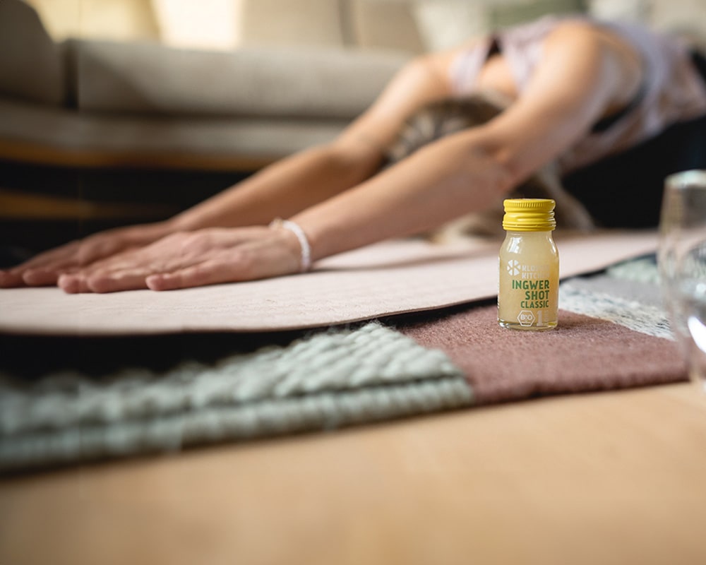 Fitness, Wellness und BGM bildlich dargestellt: Eine Dame macht Yoga, der Ingwer Shot Classic 30 ml steht dabei vor ihr.