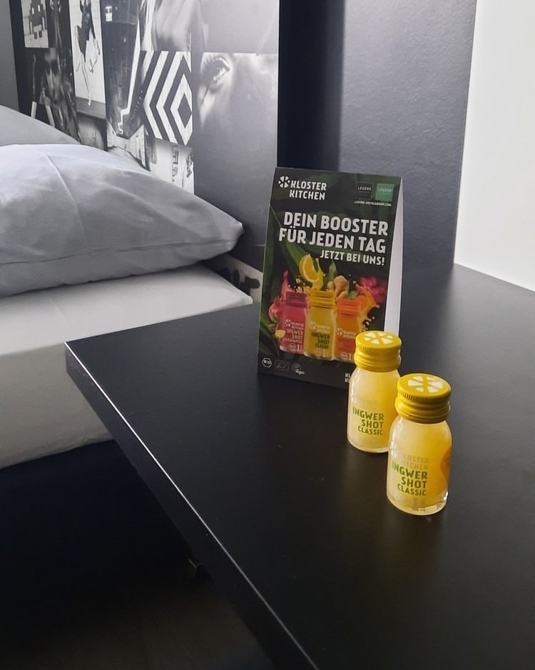 Hotel und Gastronomie bildlich dargestellt: zwei Ingwer Shot Classic 30 ml auf einem Beistelltischchen neben einem Hotelbett, mit einem Aufsteller zu den Produkten