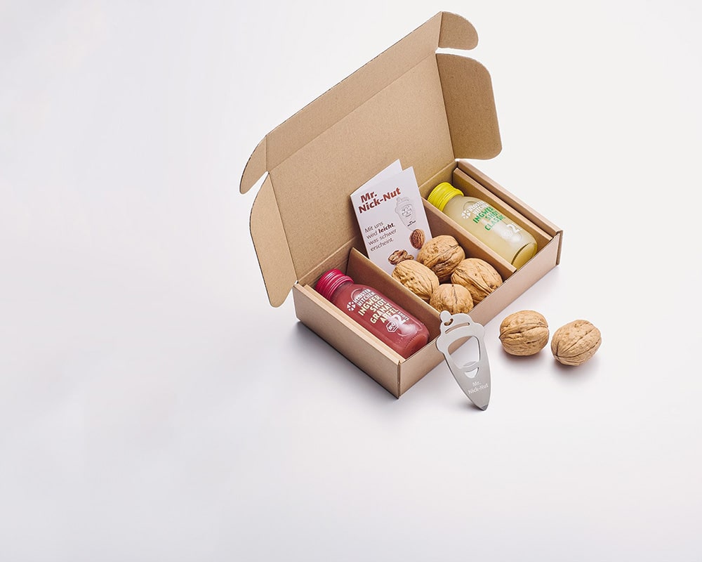 Individualisierte Artikel bildlich dargestellt: eine extra angefertigte Kartonage, in die genau zwei Ingwer Shots 60 ml hineinpassen sowie Walnüsse und ein Nussknacker / Flaschenöffner. 